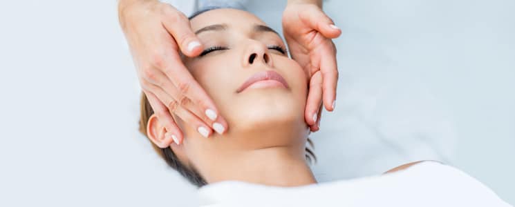 A woman lies with eyes closed as two hands rest on either side of her face during a facial