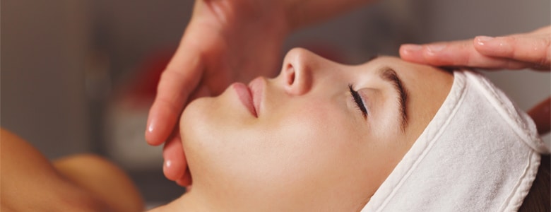 A woman in a white headband lies with eyes closed as hands touch her face during a facial