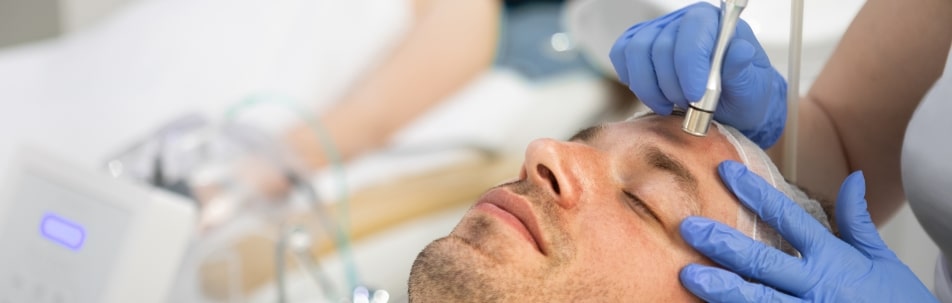 A clinician in blue gloves treats a male model’s forehead with a handheld device
